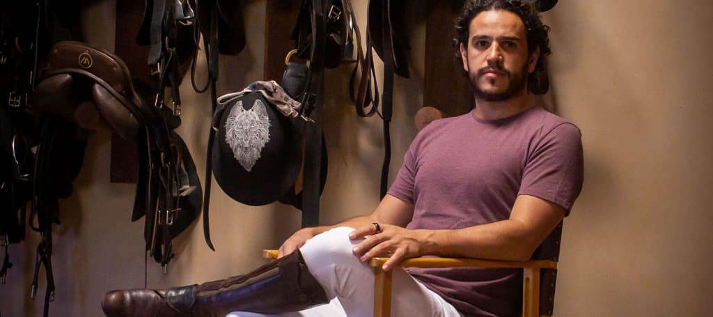 man poses in horseback riding gear in tack room for portrait