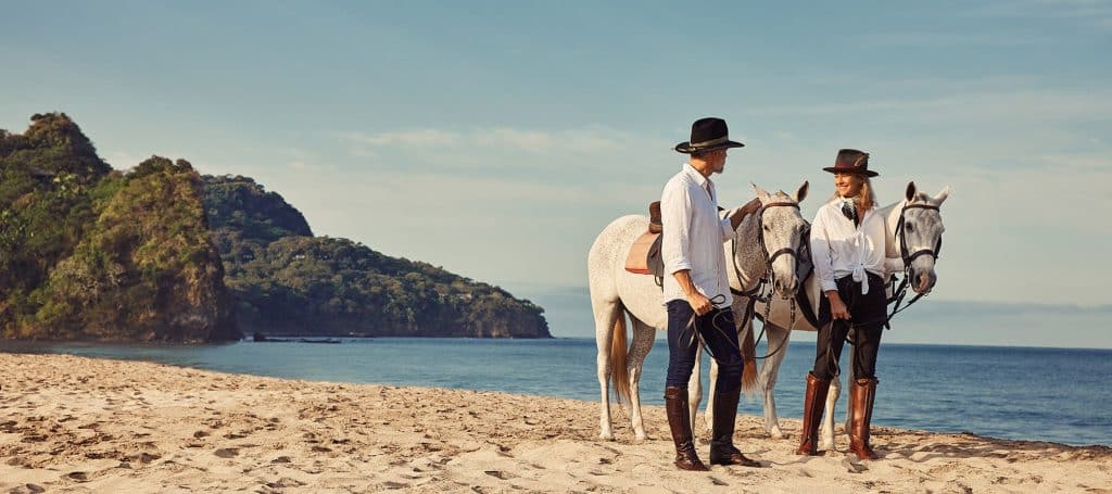 couple stands on beach with horses looking at the ocean