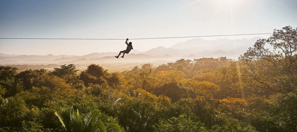 person swings high on zipline through treetops at sunset
